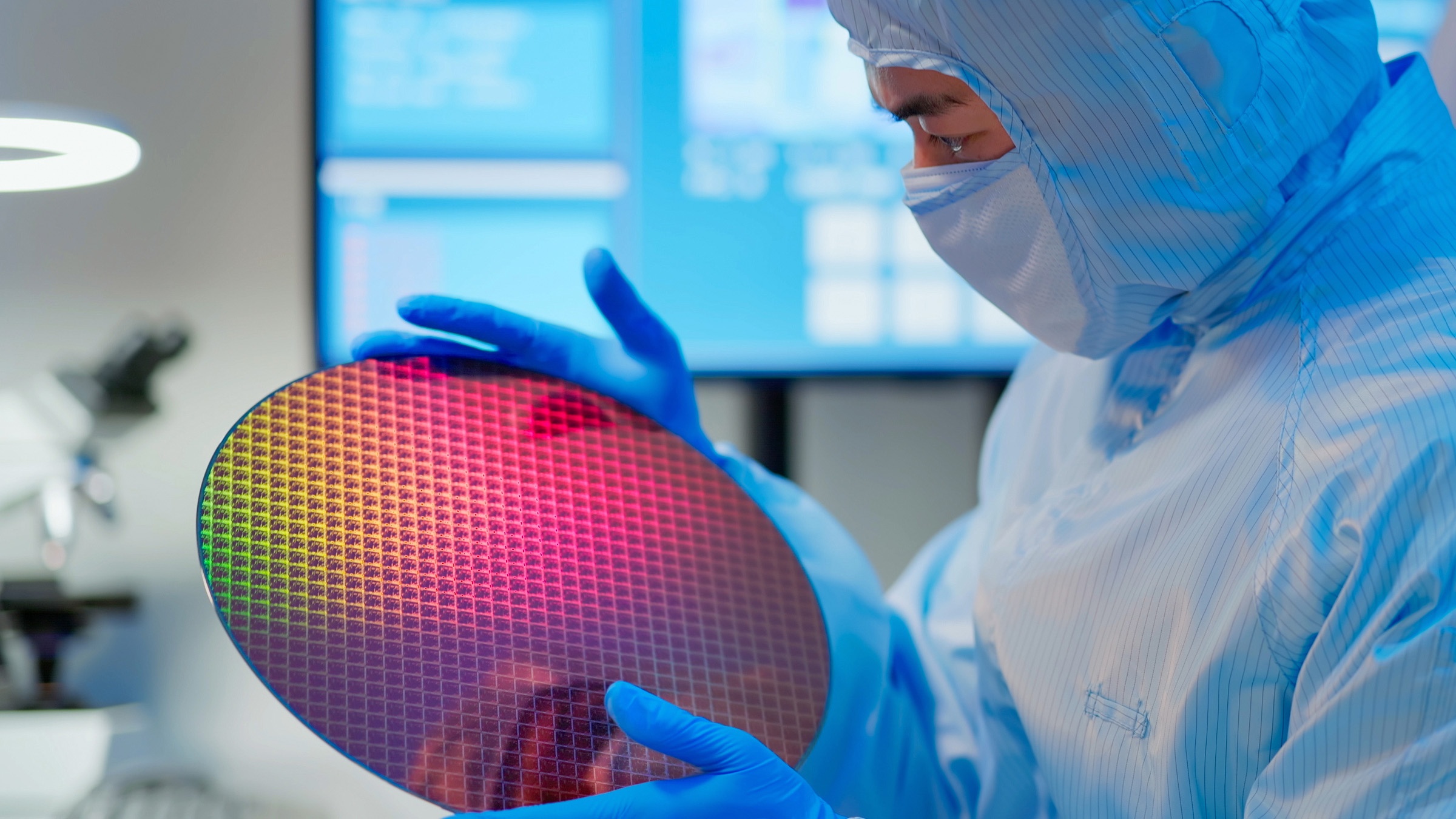 A man in a blue suit stands in a laboratory and looks at a round semiconductor plate.